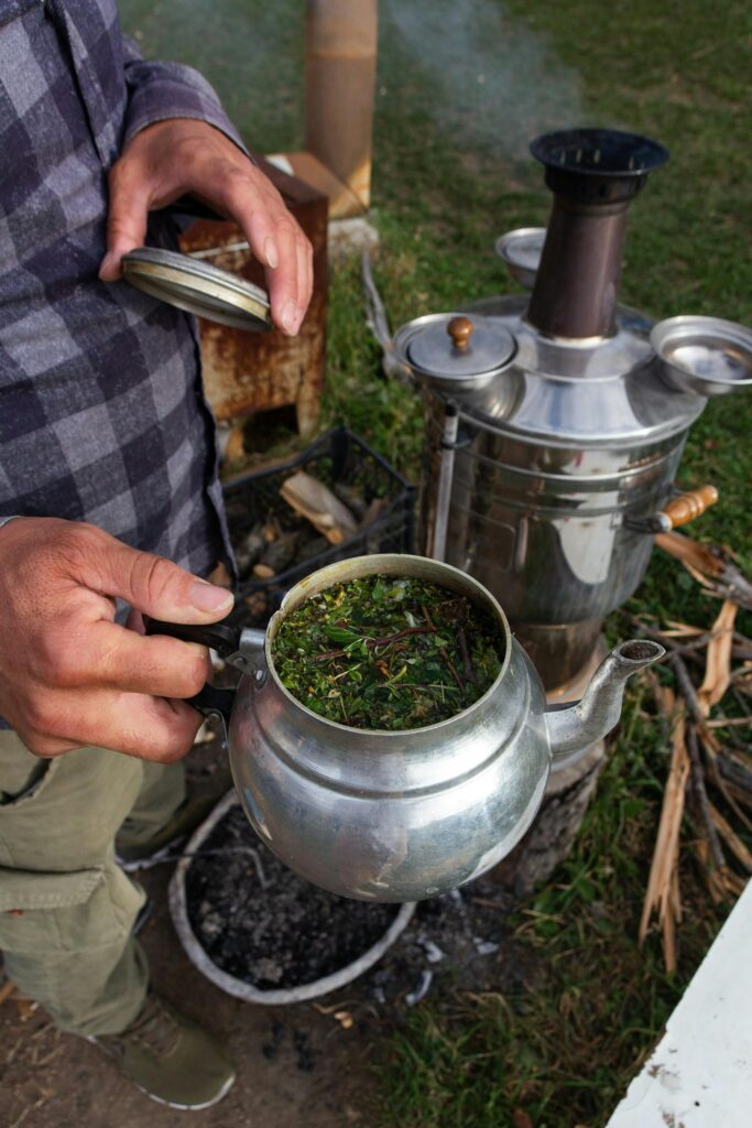 Close-up of man brewing tea outdoors with kettle and herbs.