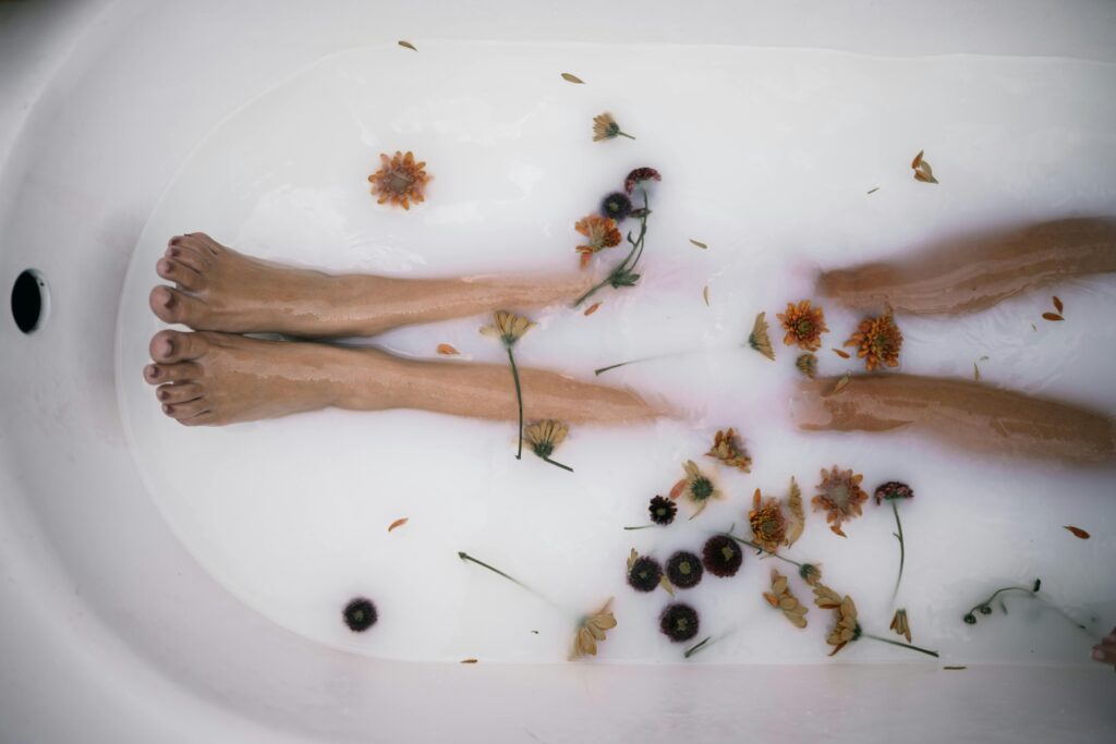 A serene bath scene showcasing legs submerged in milky water with floating flowers, emphasizing relaxation and self-care.