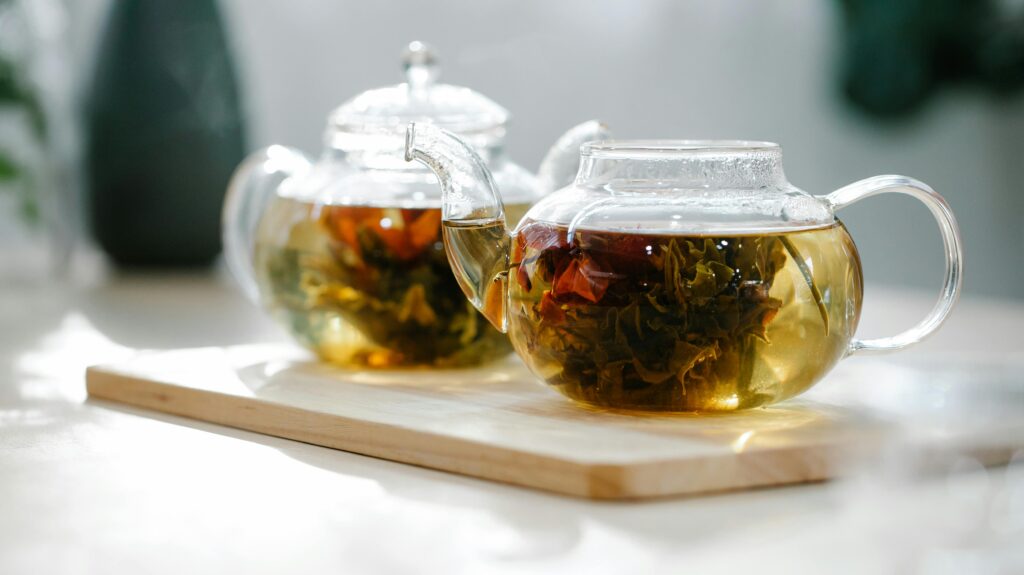 Close-up of herbal tea brewing in glass teapots placed on a wooden board, in a sunlit indoor setting.