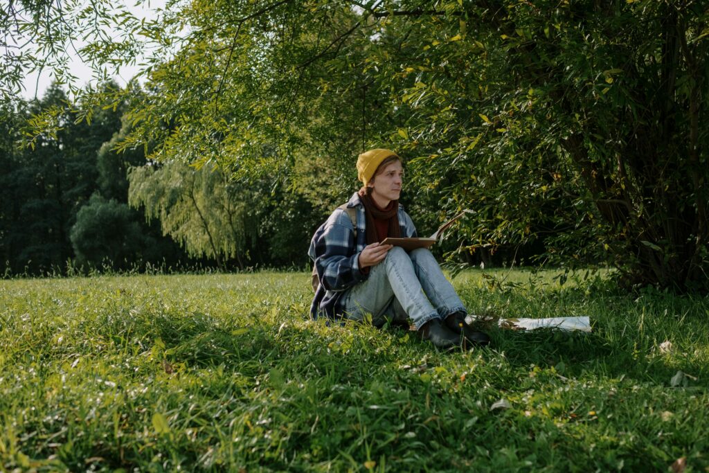 A man in a yellow beanie sits reading a book on a sunny day in a lush green park.