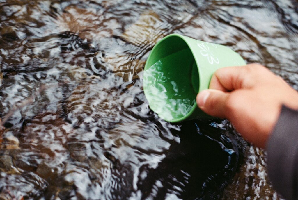 Hand collecting fresh water from a clear stream using a green cup in a natural setting.