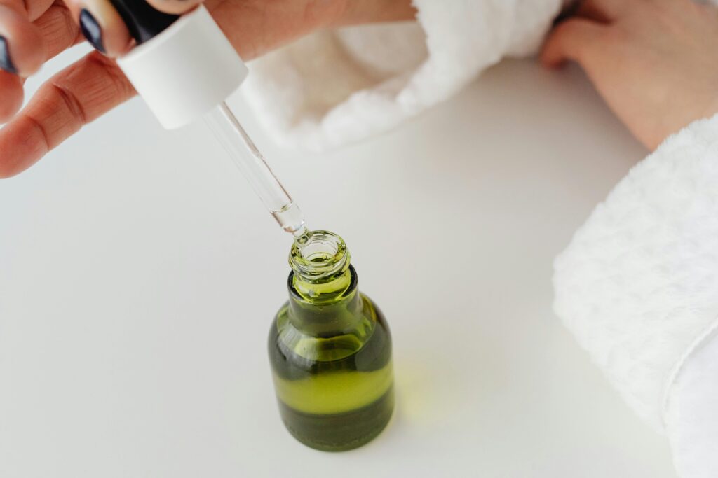 Close-up of hands with manicured nails holding a dropper over a green serum bottle.